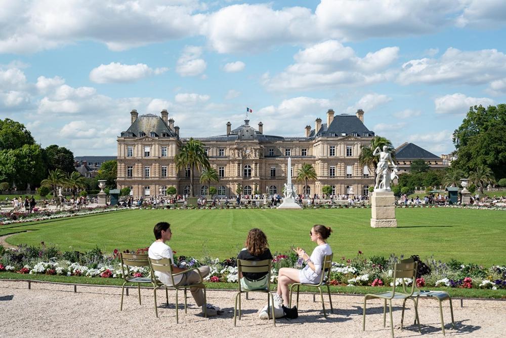 A group of people seated on chairs in front of the large building at Luxembourg Gardens, enjoying the outdoor atmosphere