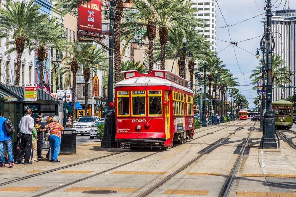 A red streetcar travels along a city street in New Orleans, surrounded by historic buildings and vibrant urban scenery.