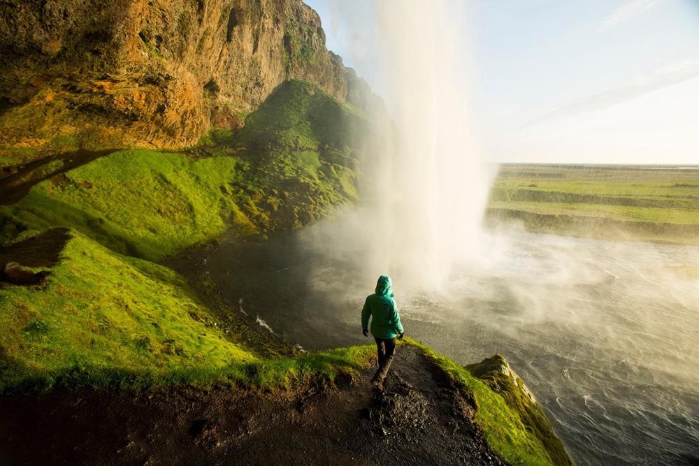 A person in a green jacket stands before a majestic geyser along Iceland's scenic Ring Road.