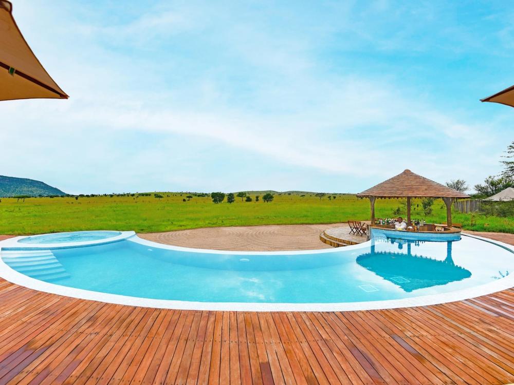  A serene infinity pool with wooden decking and umbrellas, set against a grassy field at One Nature Nyaruswiga Serengeti