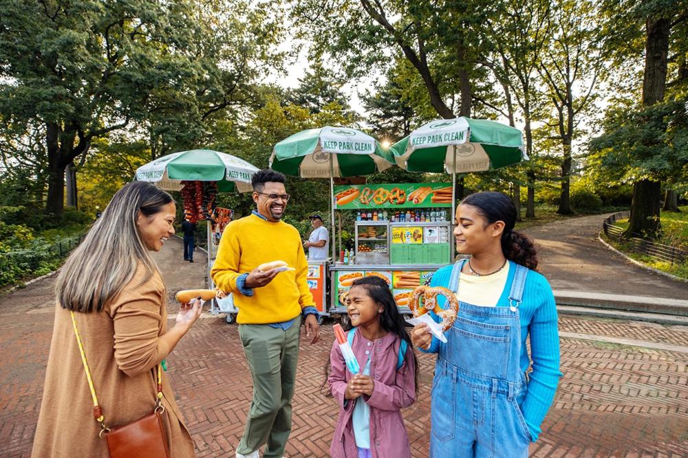 A family stands together in front of a hot dog stand, enjoying a vibrant day outdoors in NYC