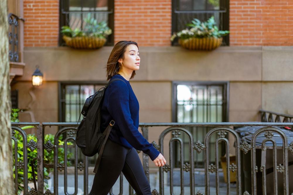 A woman in a blue sweater and wearing a black backpack walks down the street