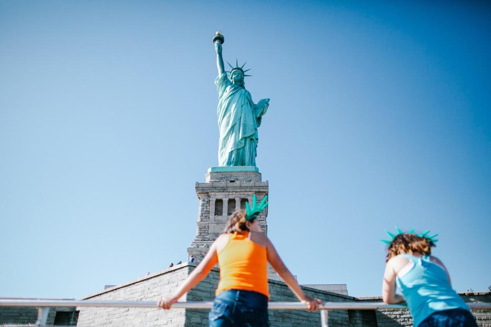 Two individuals stand in front of the Statue of Liberty, showcasing the iconic monument against a clear sky