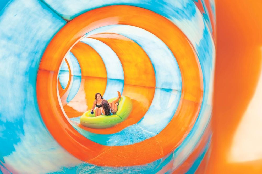 Two people joyfully descend an orange and blue water slide, splashing int the water