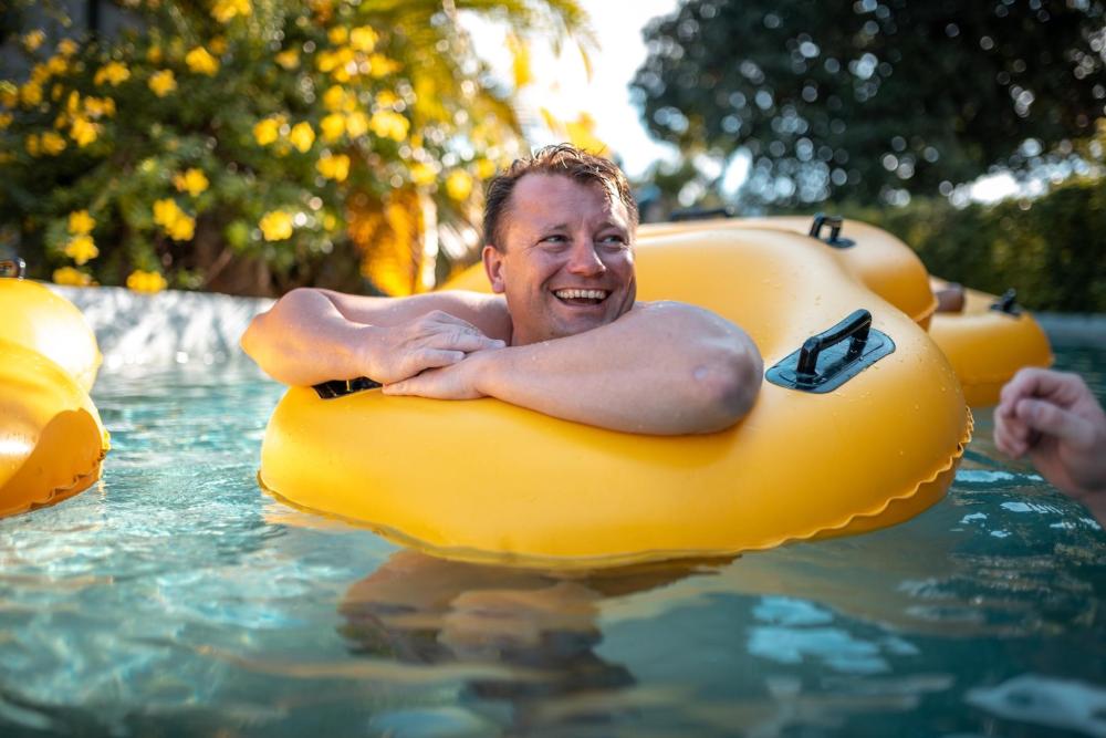 A man laughs in a pool, floating on an inflatable yellow rubber ring under the sun