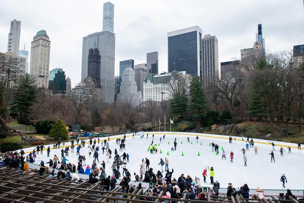 A vibrant city ice rink in New York is filled with people skating joyfully under bright lights and a cloudy sky.