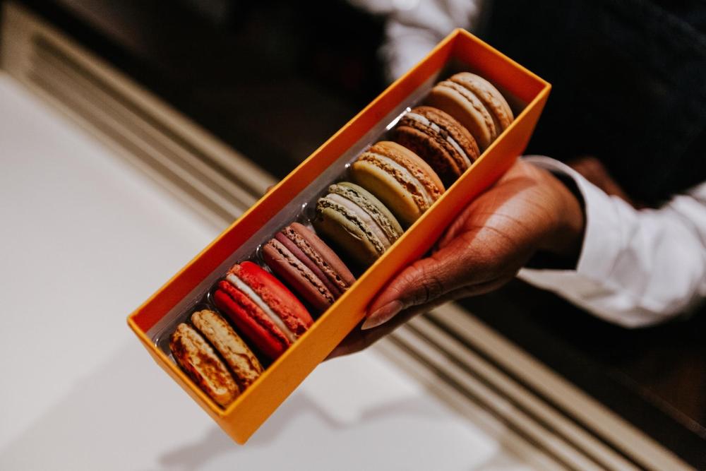 A person in Paris holds an orange box filled with an assortment of colorful macarons, showcasing a delightful treat