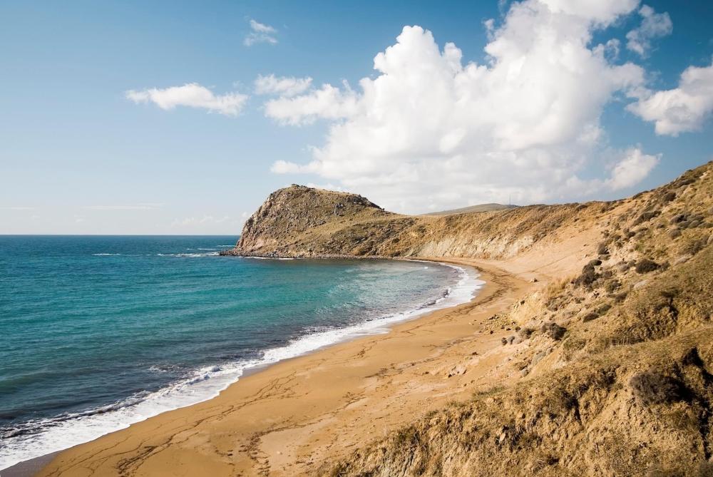 A sandy beach in Gökçeada, Turkey, featuring a rocky cliff and vibrant blue water under a cloudy sky.
