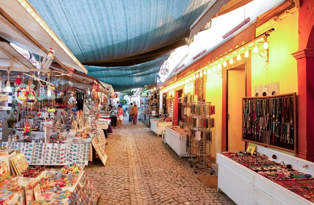 A bustling street in Cunda lined with various shops and a vibrant market, showcasing local goods and lively activity.