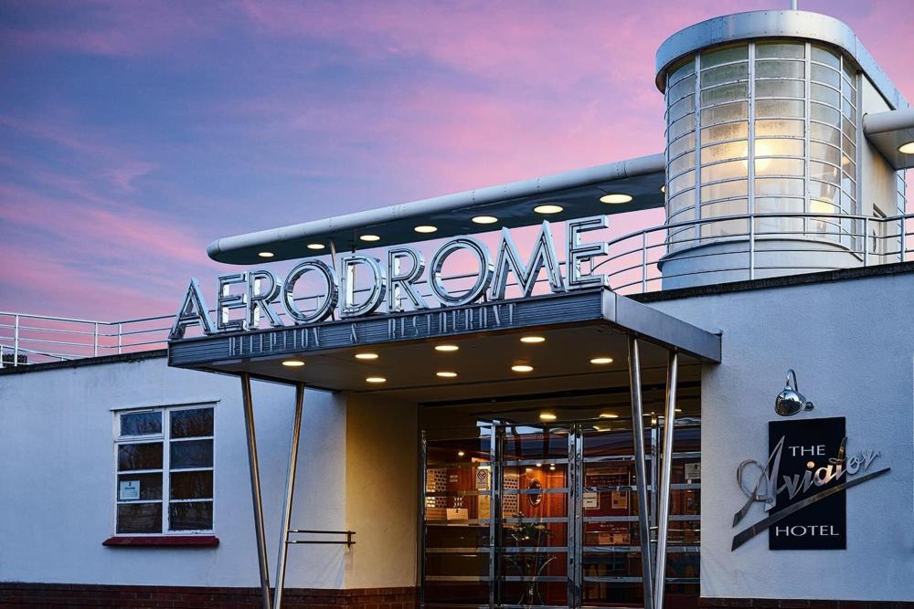 The outside of the Aviator Hotel in Sywell, UK depicted. With a purplish sky in the background, the hotel has a chrome logo which stands out boldly