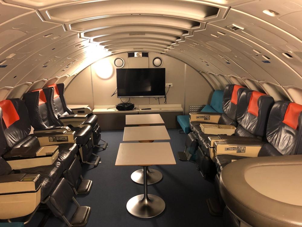 The inside of STF Jumbo Stay Stockholm is depicted. Plane seats are positioned to face one another with a flat-screen TV in the centre of the room and some tables in the middle.