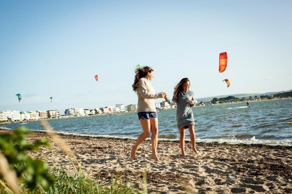 Two women run around on the beach, with a kite in the air in the background
