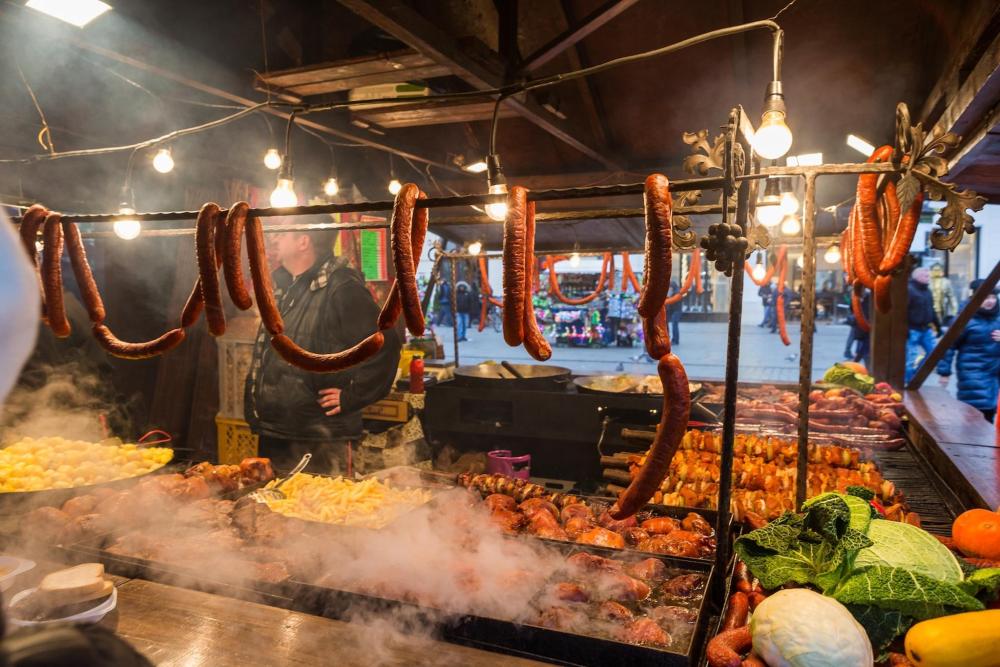 A man grilling kiełbasa krakowska at a bustling market, surrounded by fresh produce and eager customers.