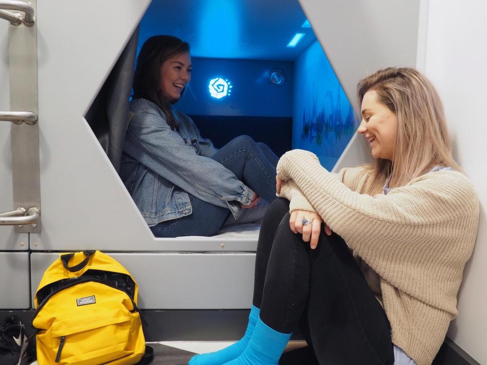 Two women sit in and beside a capsule hotel room in St. Christopher's Inn Village with a blue light in the background.