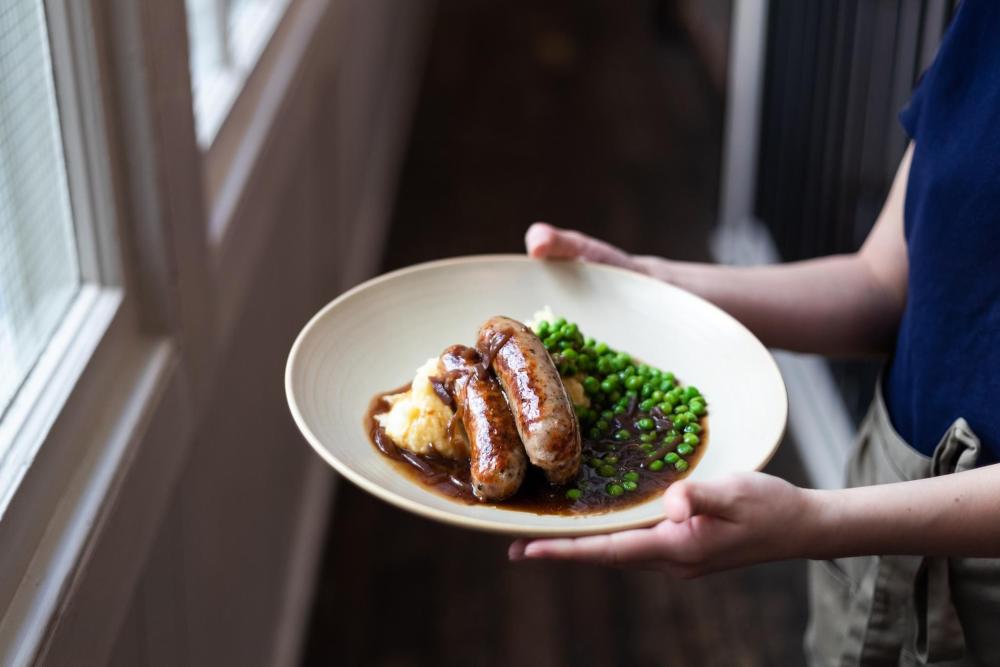 A plate of bangers and mash features, with some peas on the side and a generous serving of gravy
