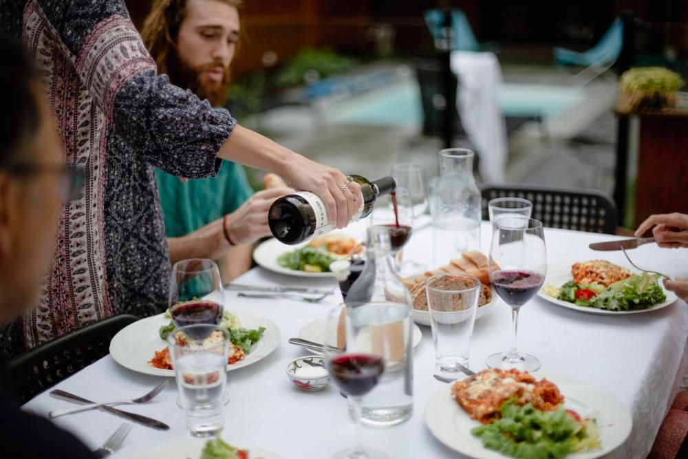 Some people are sat at a table outside. Food is on the table, and glasses of red wine are flowing.