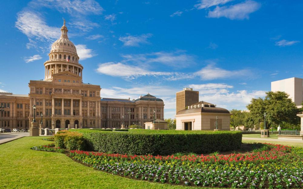 Greenery is depicted in the foreground with Texas Capitol in the background on a sunny day, with a few clouds in the sky