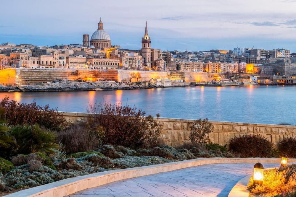 The impressive landscape of Valetta is depicted, the sun is slowly setting and the lights of the city landscape is lighting up the scene