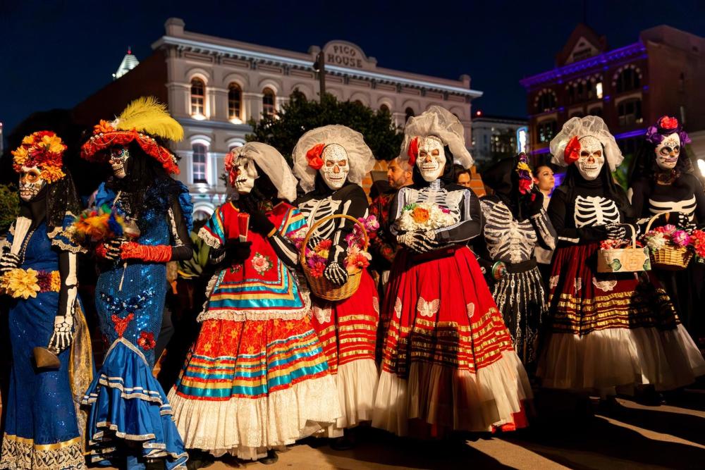 A group dressed in elaborate, colourful Día de los Muertos costumes in Mexico. Baskets of flowers are being held by the group wearing skull masks.