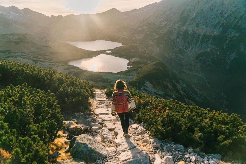 A woman with her back turned to the camera venturing through the Tatra Mountains, with some sun hitting lakes in the background