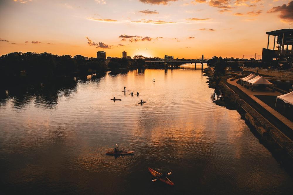 People enjoying kayaking on the river in Waco at sunset, with the sky illuminated in warm hues of orange and pink.