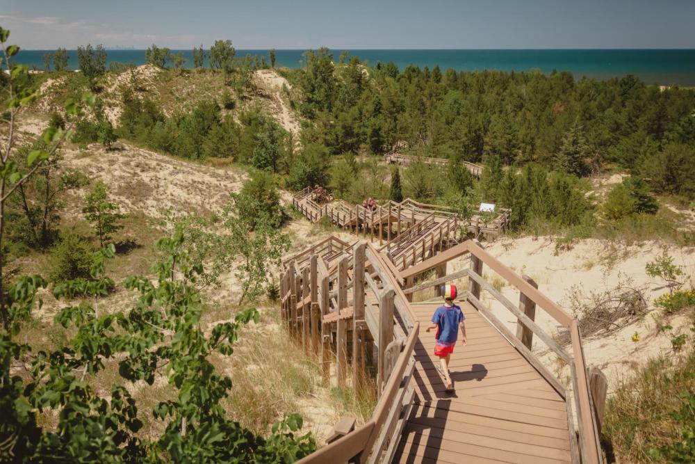 A boy strolls along a wooden boardwalk at Indiana Dunes National Park, overlooking a sandy beach and tranquil waters.