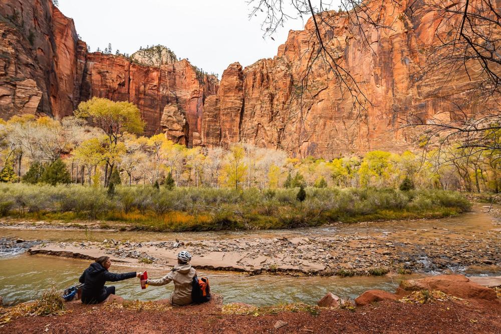 Two individuals seated by a river, with a majestic mountain backdrop in Zion National Park, Utah.