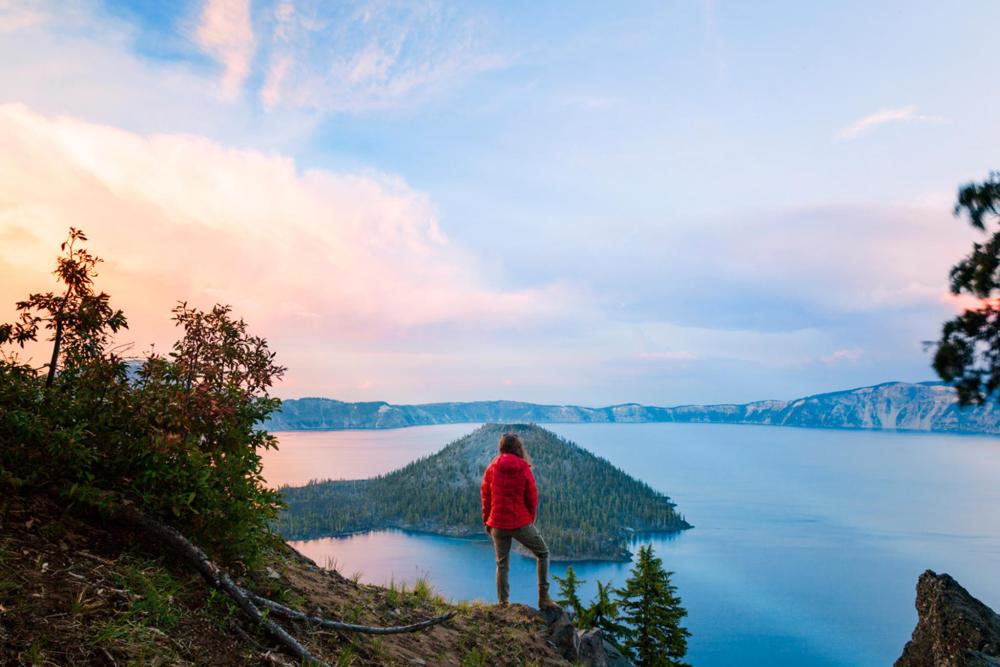  A person stands atop Crater Lake in Oregon, silhouetted against a vibrant sunset sky.