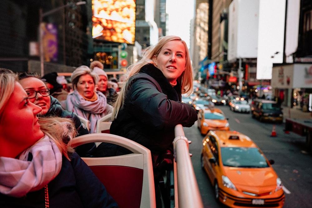 Tourists gather on a lively NYC bus tour, taking in the sights of the city. Some yellow cabs are in the background.