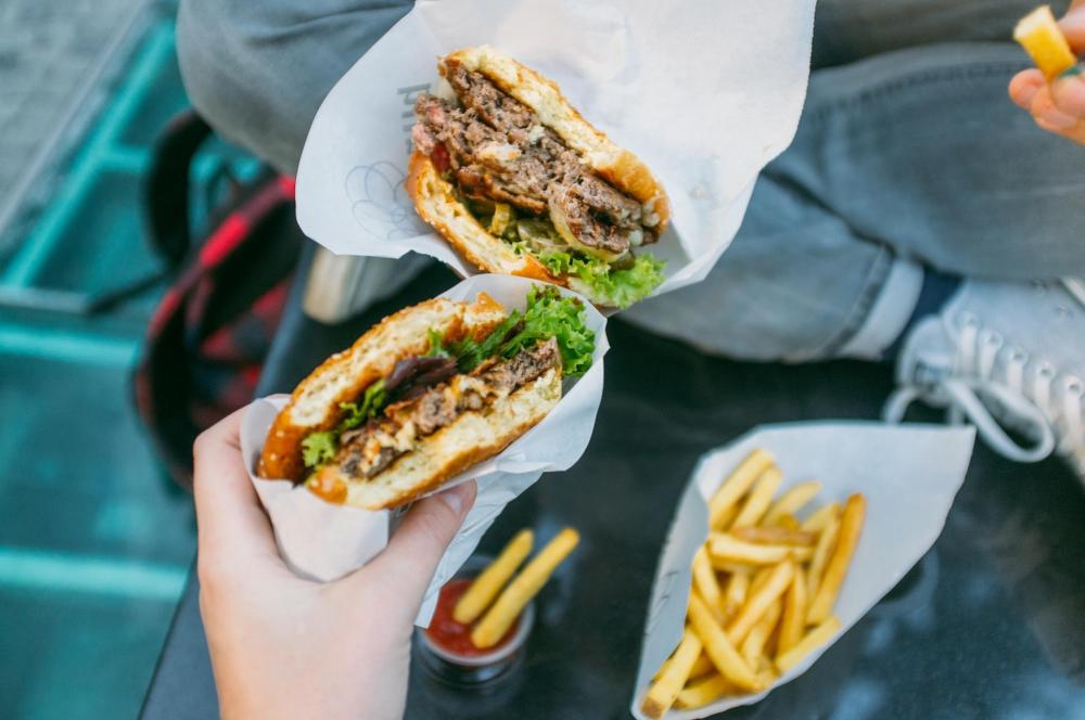 Two people hold a burger with pickles and a side of French fries