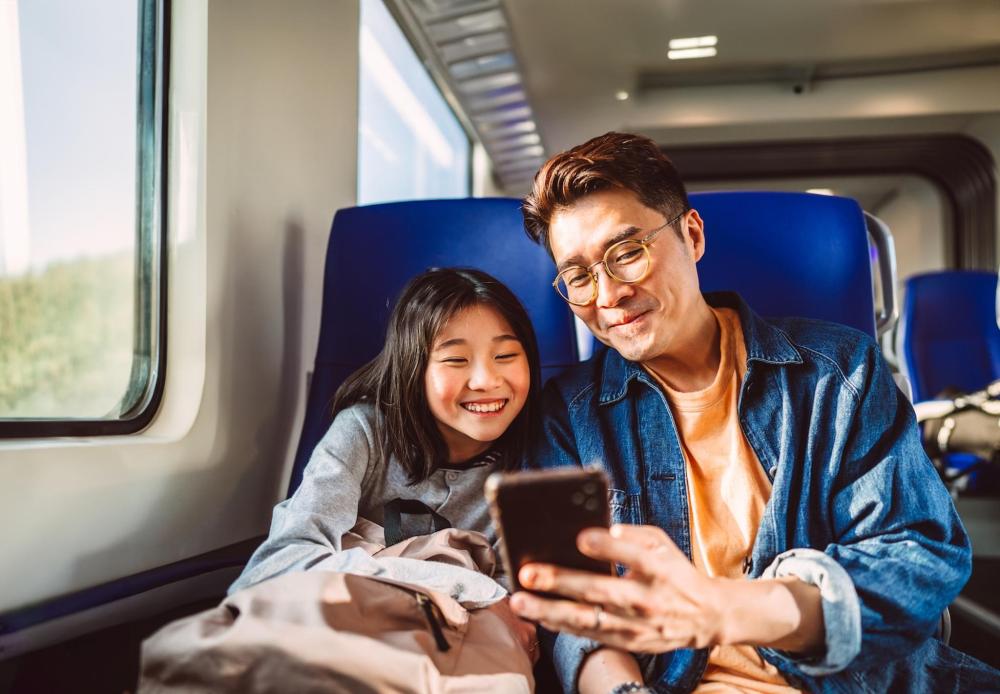 Un padre e una figlia usano il telefono insieme su un treno: sorridono con gioia, condividendo un momento.