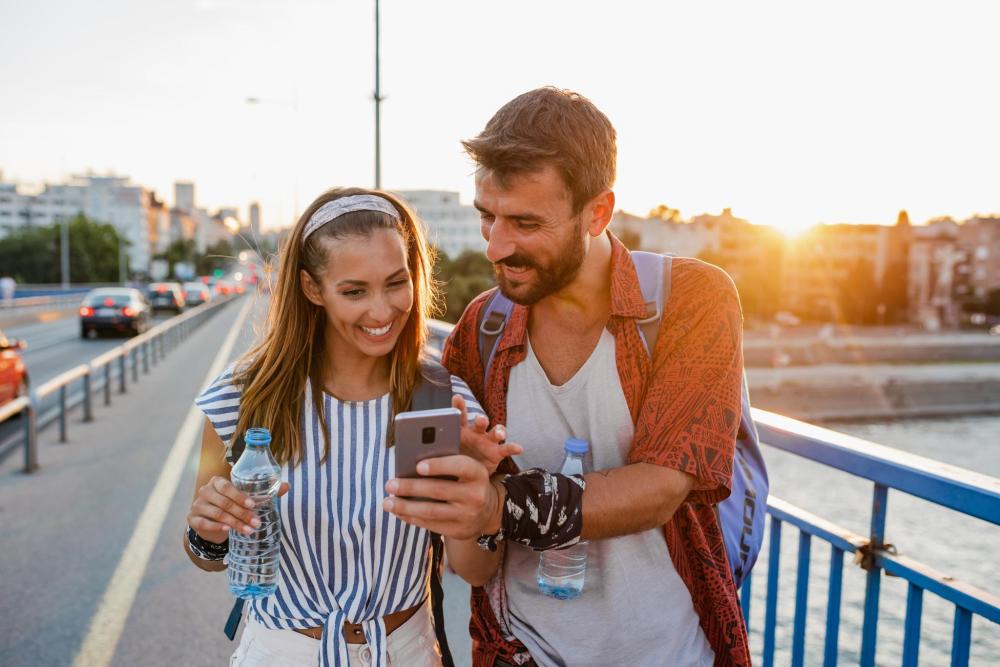 Un uomo e una donna in piedi su un ponte sorridono rivolti al cellulare. La ringhiera del ponte è visibile alle loro spalle
