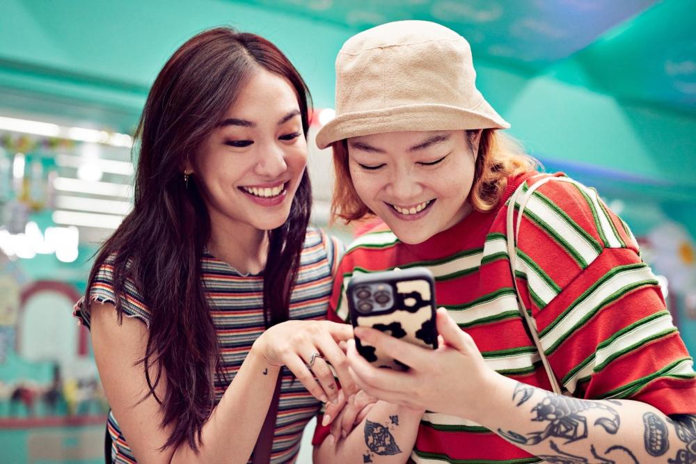 두 여성이 함께 휴대폰을 보면서 즐겁게 웃고 있다. 왼쪽 여성은 무지개 줄무늬 상의를 입었고, 오른쪽 여성은 빨간색, 흰색, 초록색 줄무늬 상의를 입고 있다.