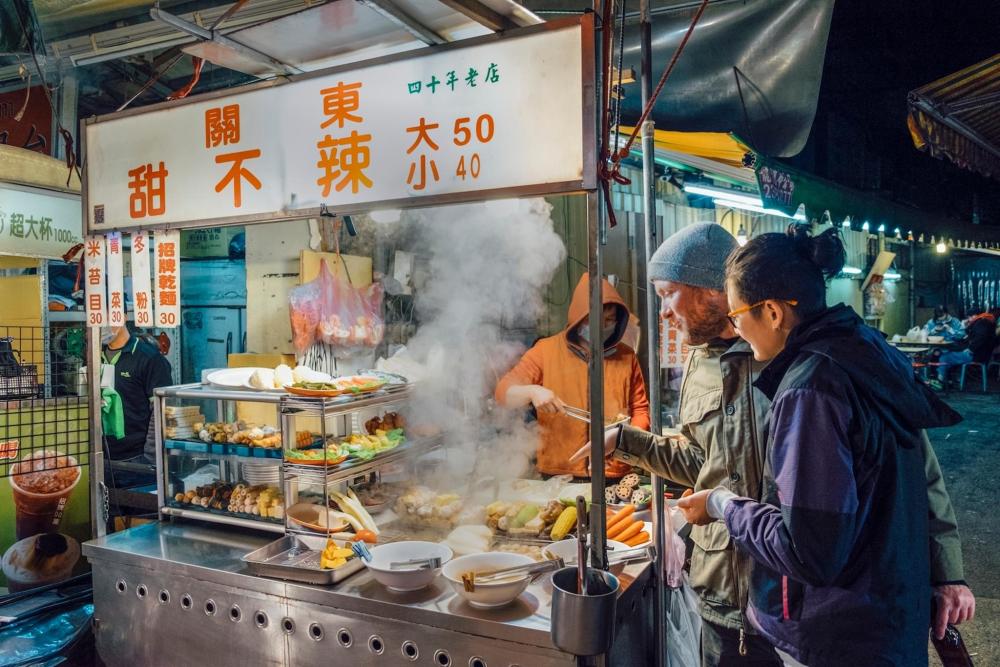 A man and woman stand in front of a food stand at a vibrant Taiwanese night market, surrounded by colourful stalls