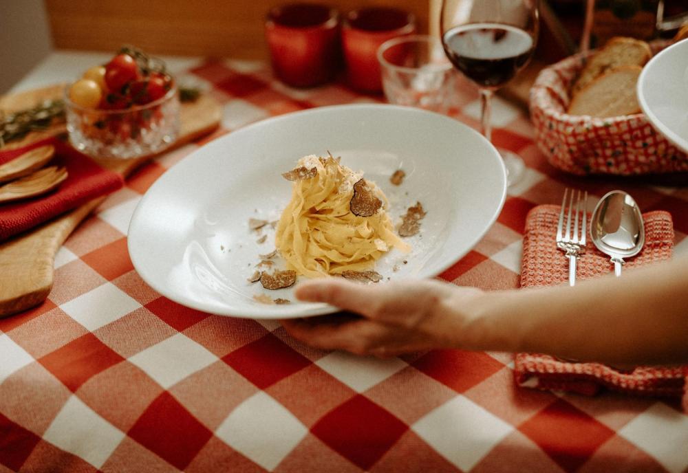 Una persona sirve un plato de pasta con trufa en una mesa con un mantel de cuadros, reflejando una comida tradicional italiana en un ambiente acogedor
