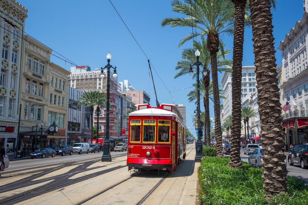 A red streetcar travels along a street in New Orleans, surrounded by historic buildings and greenery