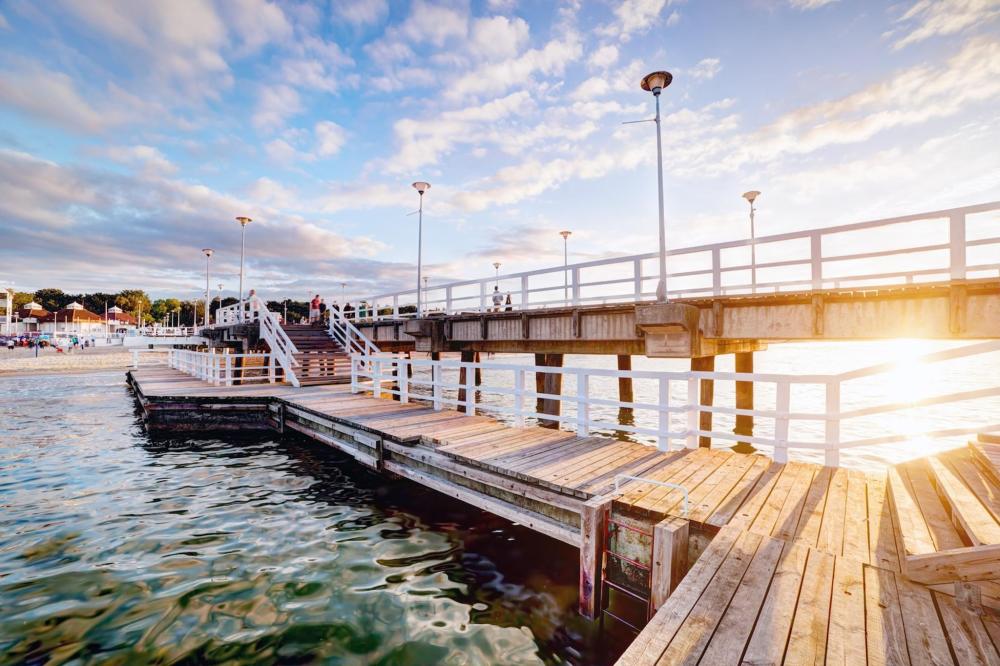 A wooden bridge spans over water at a pier in Łeba, surrounded by a serene natural landscape – golden sands can be seen in the background
