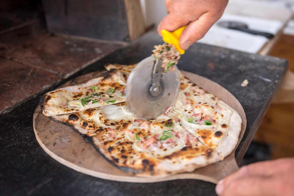A person slices a tarte flambée topped with onions, bacon and sour cream on a wooden cutting board