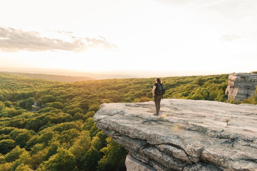 A person stands on a rock, gazing over the Catskill Mountains forest at sunset, with golden hues illuminating the sky.