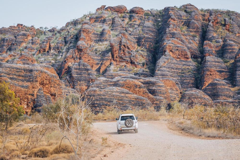 A white SUV drives along a dirt road with a rocky mountain backdrop in Australia
