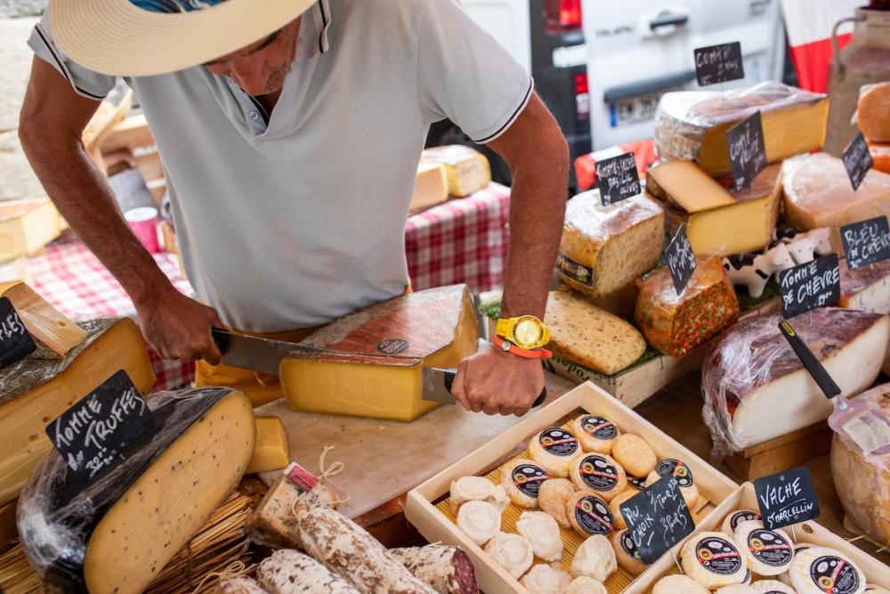A man wearing a beige hat cuts cheese in a bustling food market, surrounded by colorful produce