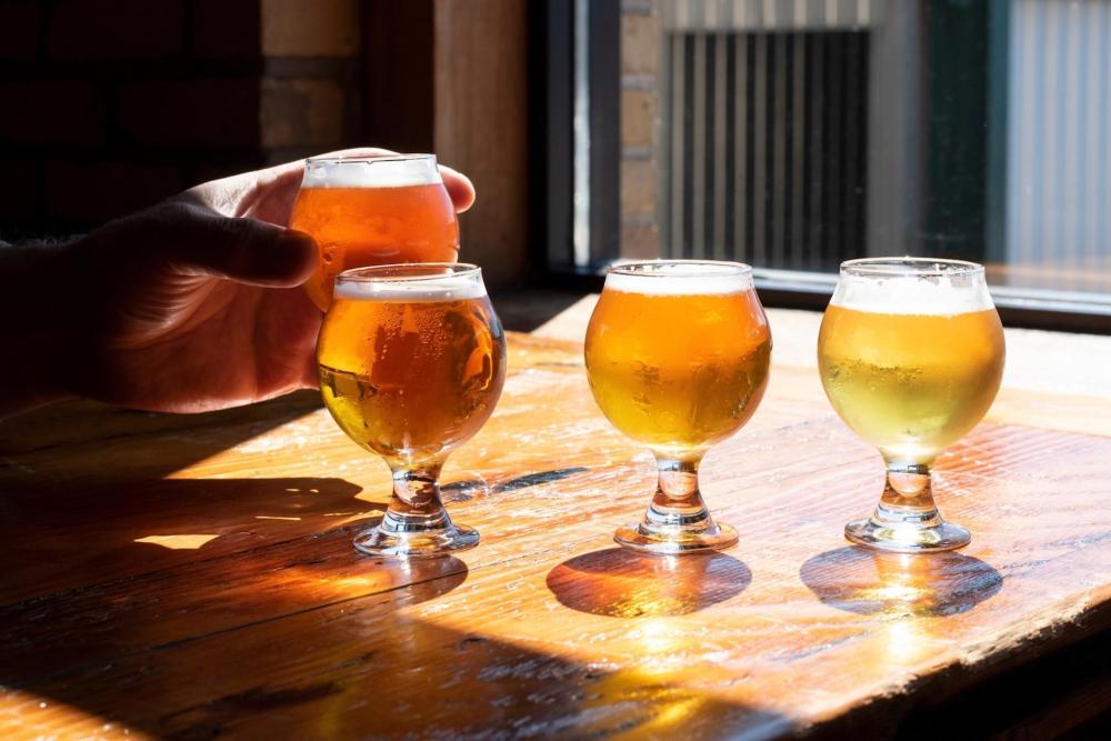 A wooden table with four small glasses of beer at a craft brewery – the sunshine from the window shines through the drink giving it a vivid appearance