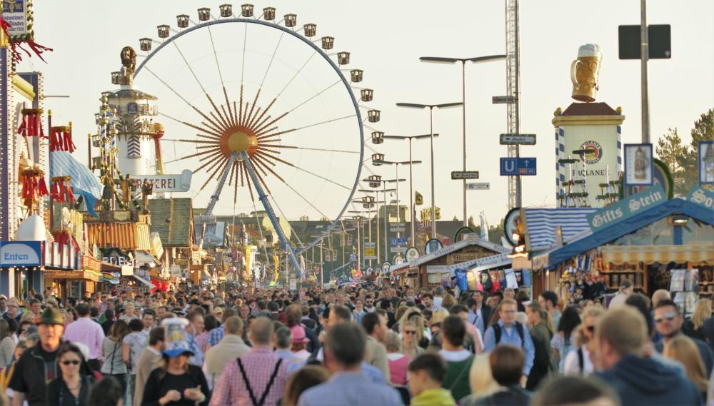 A bustling crowd at Oktoberfest walking past a colourful ferris wheel amidst decorations and food stalls