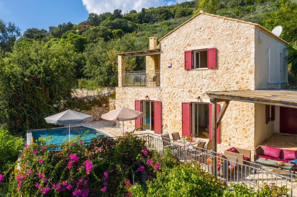 Die Villa Zoe in Griechenland verfügt über einen malerischen Pool und einen Patio inmitten von grüner Landschaft. Das Gebäude besteht aus Steinmauern mit roten Türen und Fensterläden.