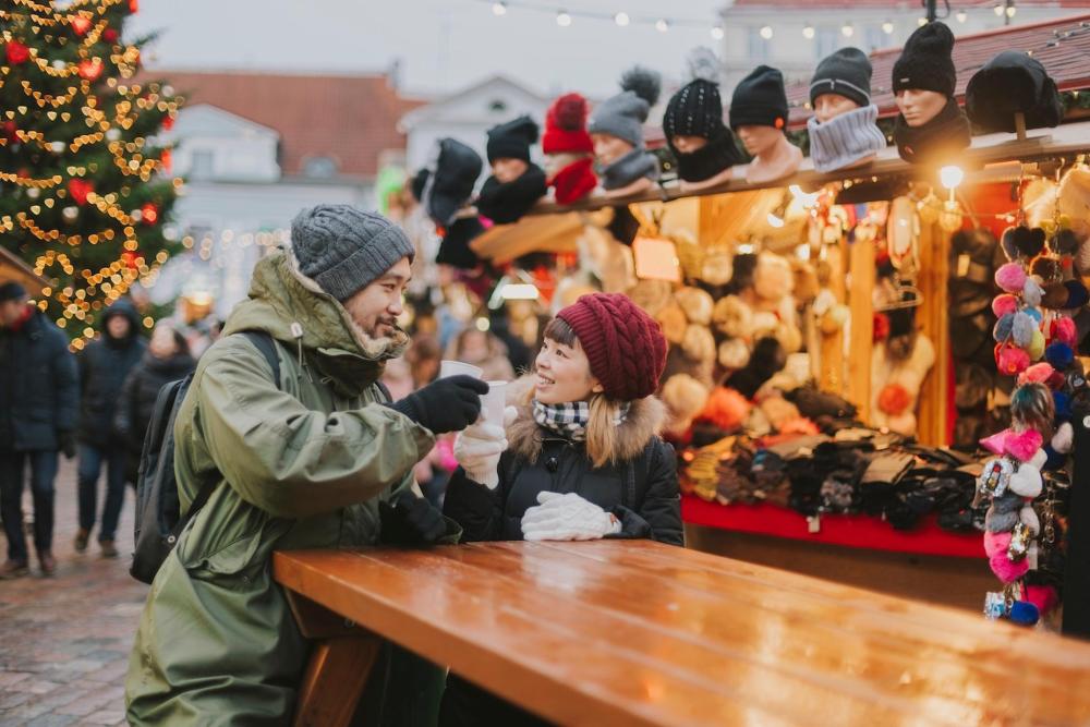 A man and woman enjoy mulled wine together at a festive Christmas market, surrounded by holiday decorations and lights