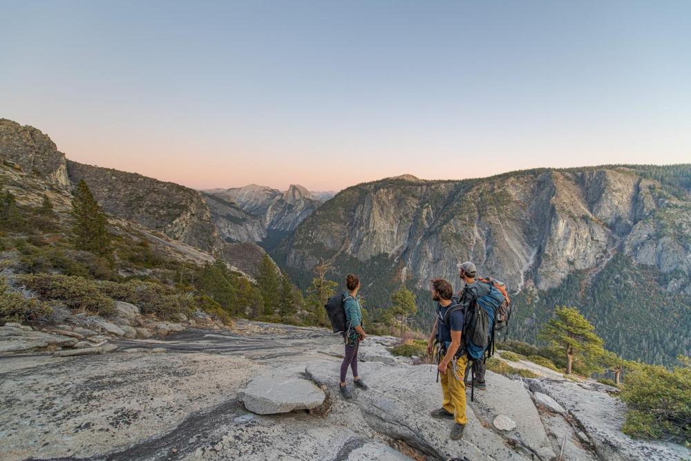 Three hikers with backpacks stand on a rock, gazing at the expansive valley in Yosemite National Park, California.