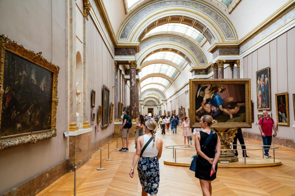 People looking at the art inside of the Louvre Museum. In the foreground there are two women walking away from the camera, with their backs turned admiring a piece of art.