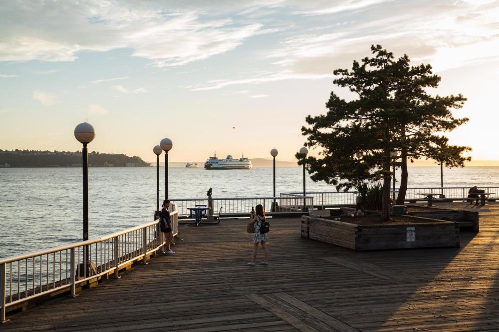 The waterfront at Seattle. Two people are depicted on the waterfront, one taking a photo of the other with a boat on the water in the distance.