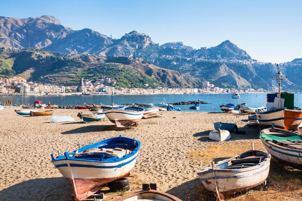 A beach in Sicily is depicted, with a few boats on the sand