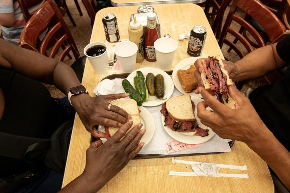 Two individuals sat at a table with Reuben sandwiches accompanied pickles, condiments and drinks at a table - engaging in a pleasant meal together